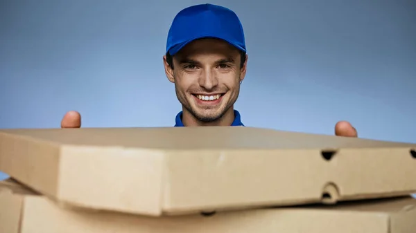 Веселий чоловік доставки дивиться на камеру, тримаючи коробки для піци на розмитому передньому плані ізольовані на синьому — стокове фото