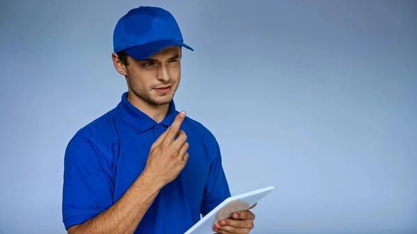 Thoughtful delivery man with digital tablet pointing with finger while looking away isolated on blue — Stock Photo