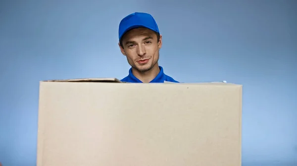Позитивний чоловік доставки дивиться на камеру, тримаючи картонну коробку ізольовано на синьому — стокове фото