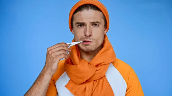 Uomo malato sconvolto in caldo cappello e sciarpa misurazione della temperatura isolato su blu — Foto stock