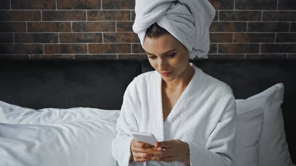 Жінка в білому рушнику дивиться на смартфон під час обміну повідомленнями в готельному номері — стокове фото