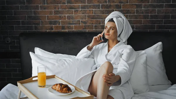 Mujer en toalla blanca y albornoz hablando en teléfono inteligente cerca de la bandeja con desayuno en la cama - foto de stock