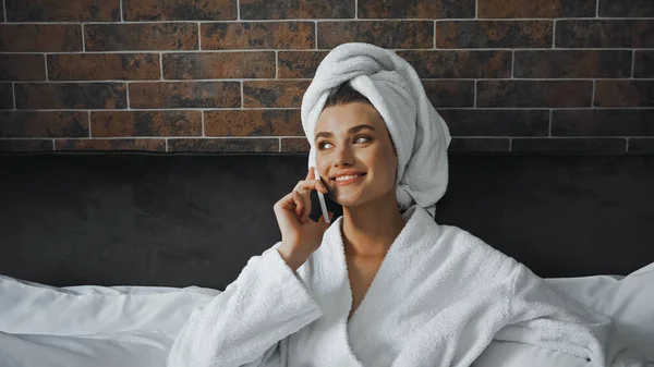 Happy woman in white towel and bathrobe talking on smartphone in hotel room — Stock Photo