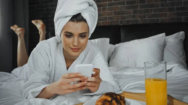 Mujer joven en toalla con teléfono inteligente cerca del desayuno en bandeja en la habitación del hotel - foto de stock