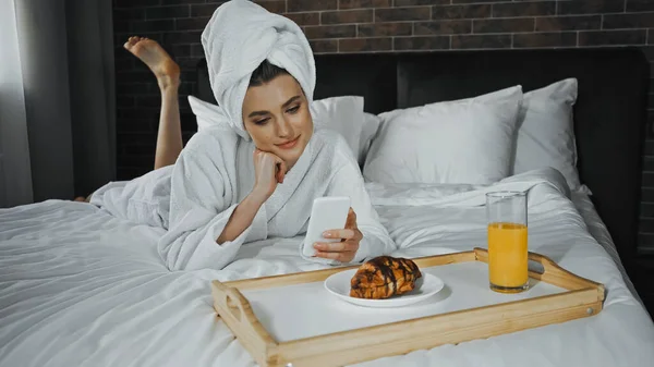 Jovem mulher em toalha e roupão de banho usando smartphone perto do café da manhã na bandeja no quarto do hotel — Fotografia de Stock