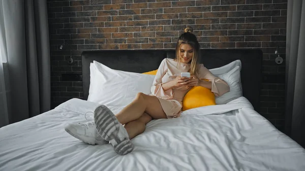 Donna allegra sorridente durante la messaggistica su smartphone in camera da letto — Foto stock