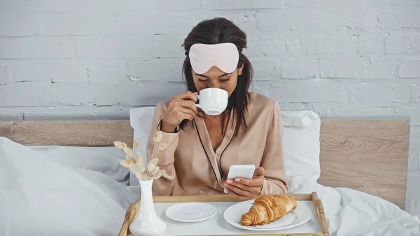 Брюнетка жінка п'є каву і обмін повідомленнями на смартфоні в ліжку — стокове фото