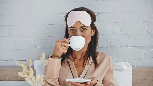 Mujer alegre con máscara para los ojos beber café y mirando a la cámara - foto de stock