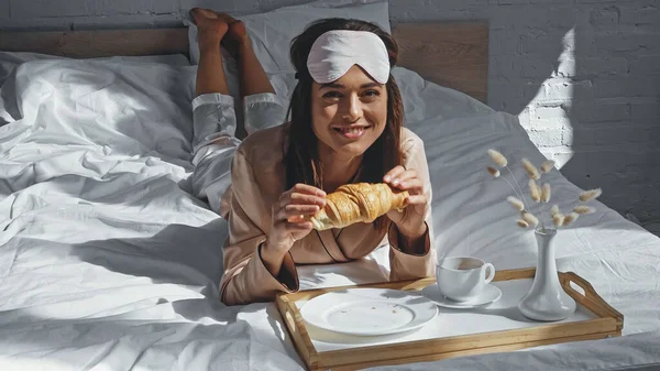 Joyful woman lying in bed and holding croissant near tray with breakfast — Stock Photo