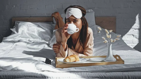 Mulher deitada na cama e segurando xícara enquanto bebe café perto da bandeja com café da manhã — Fotografia de Stock