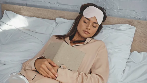 Женщина в маске глаза спит с книгой в постели — стоковое фото
