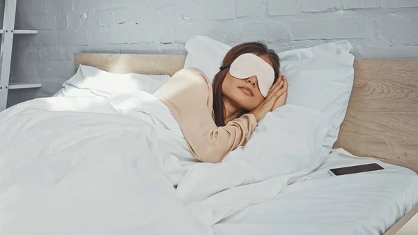 Mujer con máscara ocular durmiendo cerca de teléfono inteligente con pantalla en blanco - foto de stock