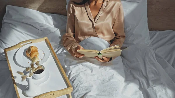 Vista recortada de la mujer leyendo libro cerca de la bandeja con sabroso desayuno - foto de stock
