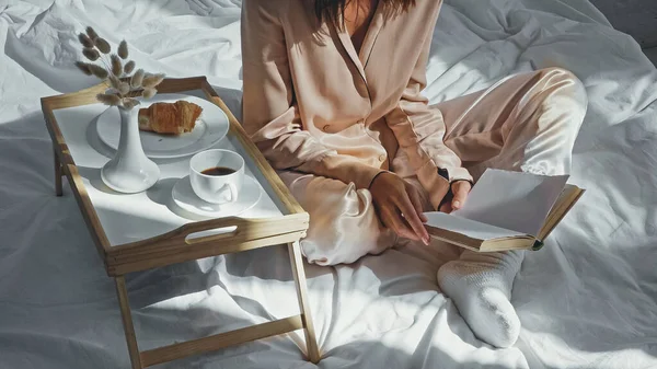 Vista ritagliata di donna con gambe incrociate libro di lettura vicino vassoio con gustosa colazione — Foto stock
