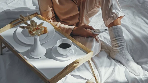 Vista cortada da mulher usando smartphone perto da bandeja com café da manhã saboroso — Fotografia de Stock