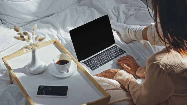 Visão de alto ângulo da mulher segurando xícara de café perto do laptop com tela em branco na cama — Fotografia de Stock