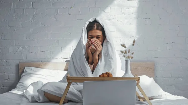 Frau trinkt warmen Tee, während sie unter einer Decke in der Nähe des Frühstückstabletts sitzt und einen Film auf dem Laptop anschaut — Stockfoto