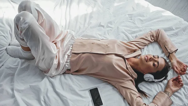 Hochwinkel-Ansicht einer glücklichen Frau in drahtlosen Kopfhörern, die auf dem Bett in der Nähe des Smartphones mit leerem Bildschirm liegt — Stockfoto