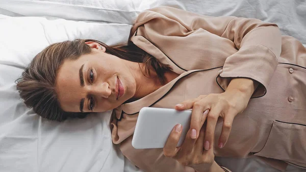 Вид сверху веселой женщины, болтающей на смартфоне, лежа в постели — стоковое фото