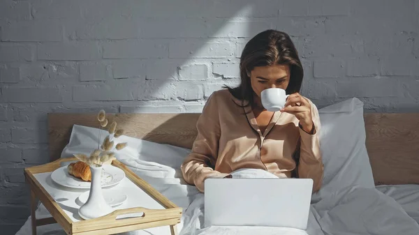 Femme regardant un film sur ordinateur portable et buvant du thé près du plateau avec petit déjeuner — Photo de stock