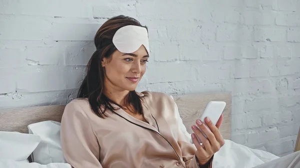 Mulher feliz em máscara de olho olhando para o smartphone no quarto — Stock Photo