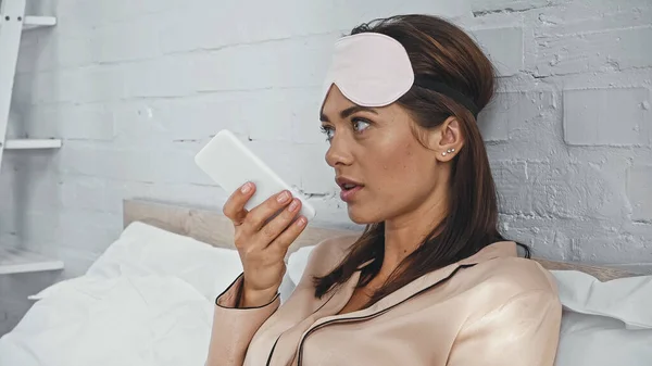 Mujer joven con máscara ocular sosteniendo teléfono inteligente y grabando mensaje de voz en el dormitorio - foto de stock
