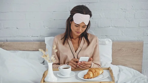 Mulher morena mensagens no smartphone perto da bandeja com café da manhã saboroso — Fotografia de Stock