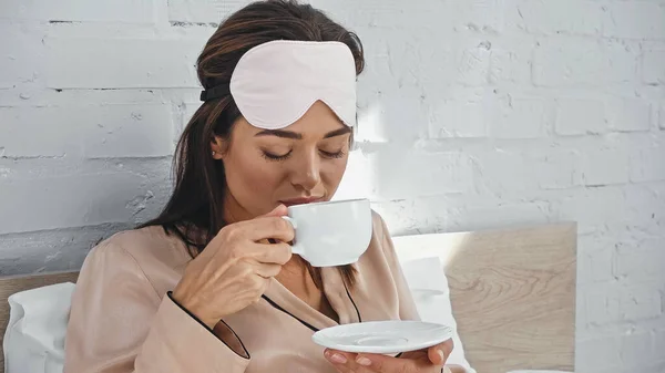 Jovem mulher em máscara olho segurando copo e pires enquanto bebe café — Fotografia de Stock