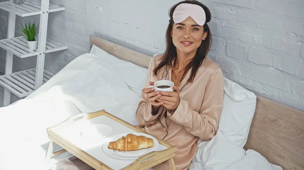 Mulher feliz segurando xícara de café perto de croissant na bandeja na cama — Fotografia de Stock