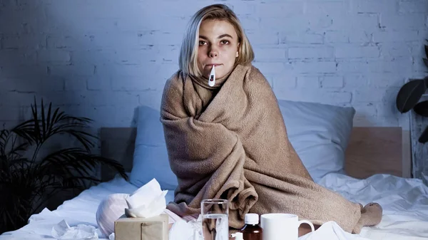 Diseased woman measuring temperature while sitting under blanket near medications — Stock Photo