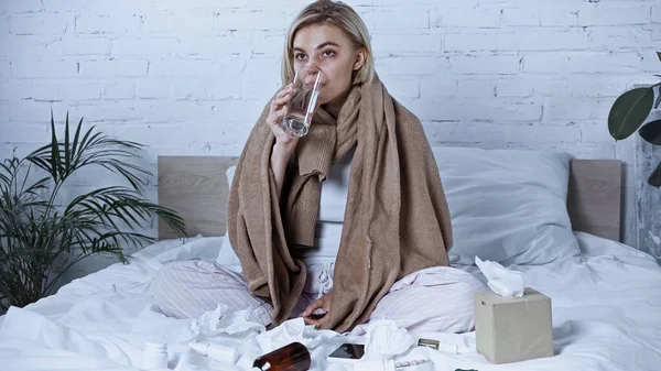 Kranke Frau trinkt Wasser, während sie in der Nähe von Medikamenten im Bett sitzt — Stockfoto