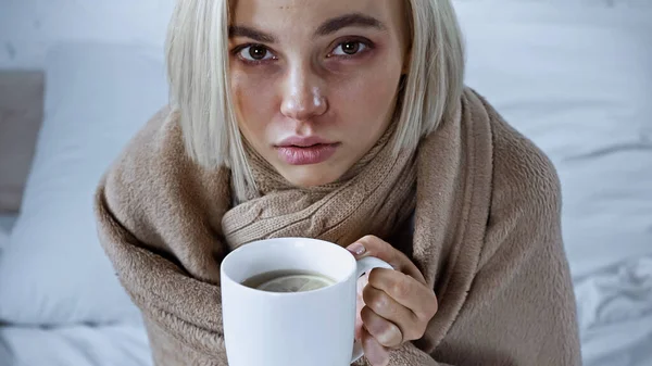 Sick woman, wrapped in blanket, holding cup of warm tea in bedroom — Foto stock