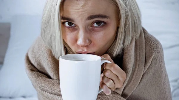 Ill woman looking at camera while drinking warm beverage in bedroom — Stock Photo
