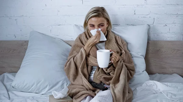 Хвора жінка чхає в паперову серветку під час перегляду телевізора в спальні з чашкою чаю — стокове фото
