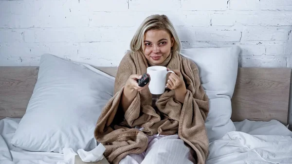 Хвора, усміхнена жінка натискає телевізійні канали, тримаючи чашку чаю в спальні — стокове фото