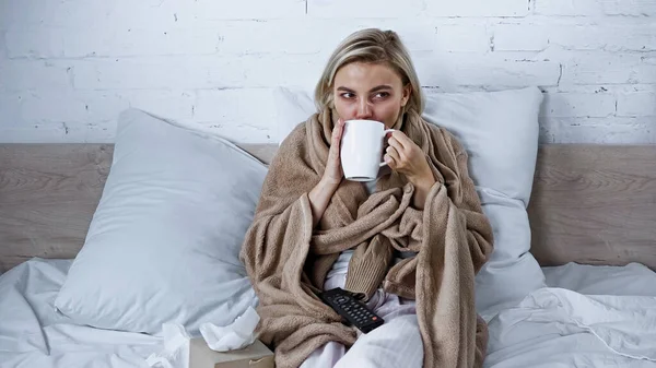 Diseased woman drinking warm beverage while watching tv in bedroom — Stock Photo