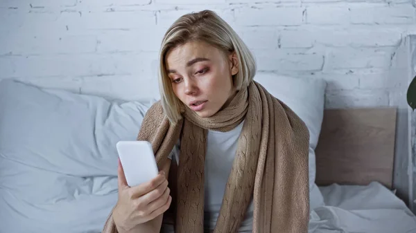 Засмучена хвора жінка тримає смартфон, сидячи під теплою ковдрою в спальні — стокове фото