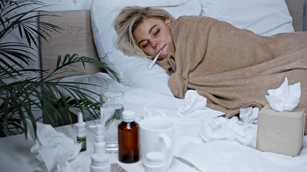 Sick woman measuring temperature in bed near napkins and medications on blurred foreground — Stock Photo