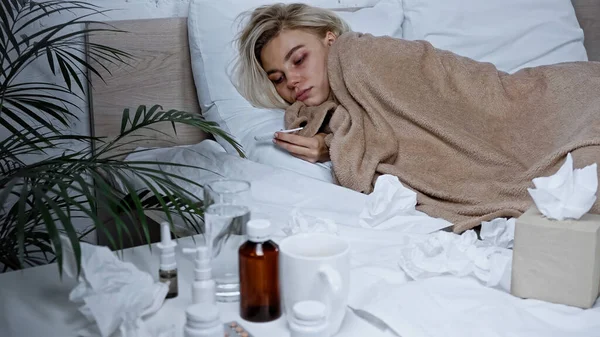 Sick woman holding thermometer while lying in bed near medications on blurred foreground — Stock Photo