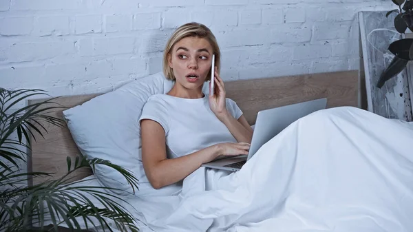 Блондинка фрилансер с помощью ноутбука во время разговора на мобильном телефоне в спальне — стоковое фото