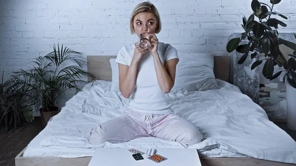 Sick woman drinking water while sitting on bed near table with medications — Stock Photo