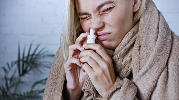 Femme malade, recouverte d'une couverture chaude, fronçant les sourcils en utilisant un spray nasal — Photo de stock