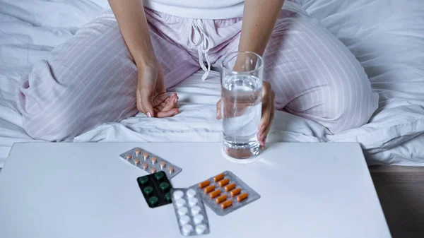 Partial view of diseased woman sitting on bed with glass of water near table with pills — Stock Photo