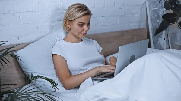 Young blonde freelancer working on laptop in bedroom — Stock Photo