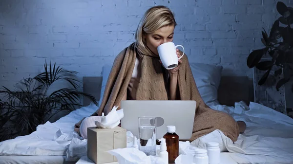 Sick freelancer drinking warm tea near laptop and medications in bedroom — Stock Photo