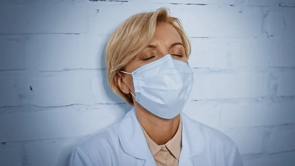 Tired doctor in medical mask standing near brick wall — Stock Photo