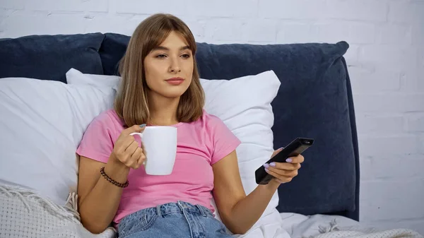 Junge Frau hält Tasse und Fernbedienung in der Hand, während sie Film im Schlafzimmer schaut — Stockfoto