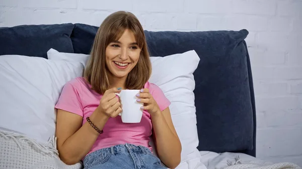 Счастливая молодая женщина держит чашку и смотрит кино в спальне — стоковое фото