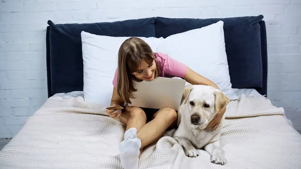 Молода жінка тримає кредитну картку біля ноутбука та собаки-качалки на ліжку — стокове фото