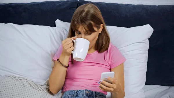 Jeune femme boire du café et en utilisant smartphone dans la chambre — Photo de stock